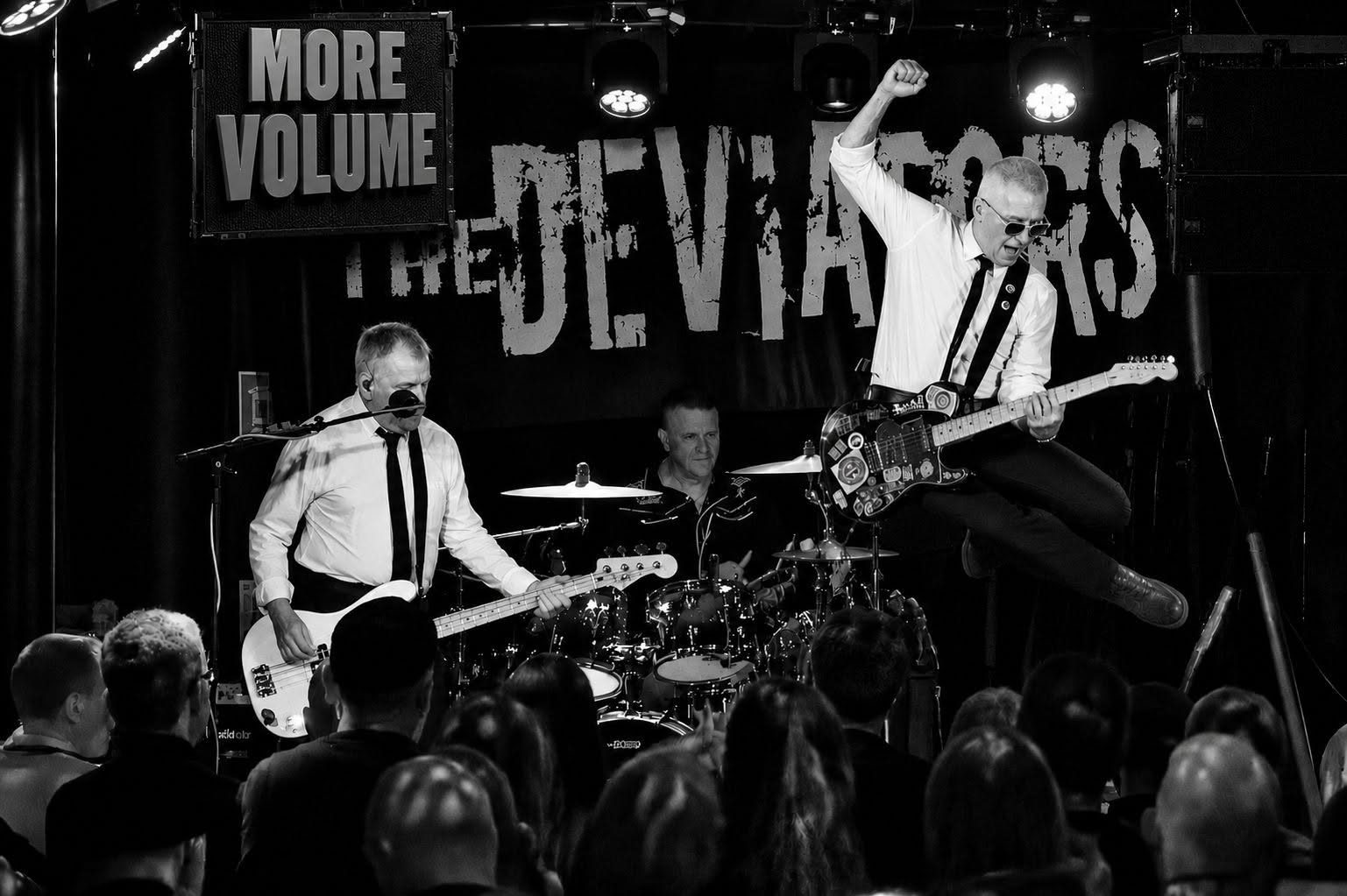 Bitzy mid-air with his guitar at The Grand Social, Bren on bass and Andy on drums under the distressed Deviators backdrop